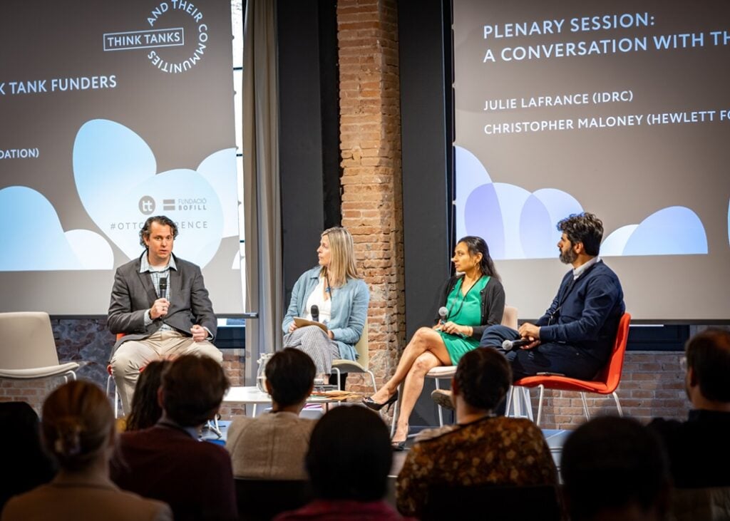 The facilitator and three think tank funders sit on the stage with microphones. In the foreground is the outline of audience members.